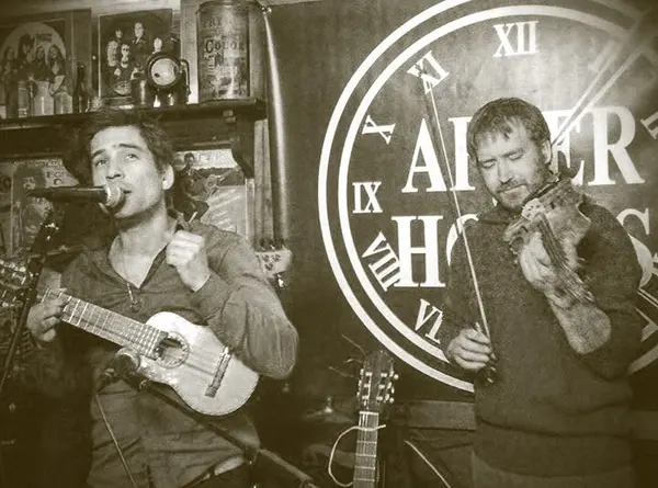 En sépia, dans un bar : un musicien chante en jouant d'un charango, l'autre joue du violon devant une grande horloge.