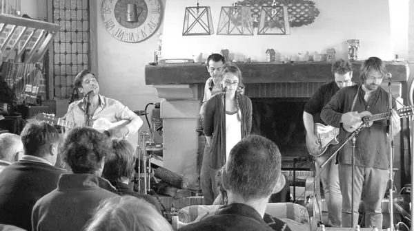 En noir et blanc, concert dans un salon devant une cheminée : guitares, chant et chœurs, le public assis tout près.