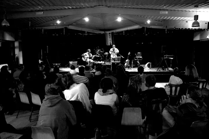 Concert sous un grand chapiteau : le public assis de dos, le duo sur la scène éclairée au fond.