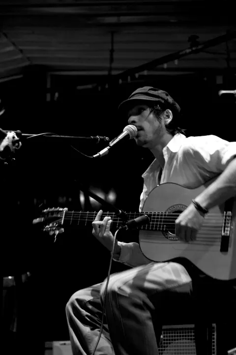 En noir et blanc, un musicien chante en jouant de la guitare, assis sur scène.
