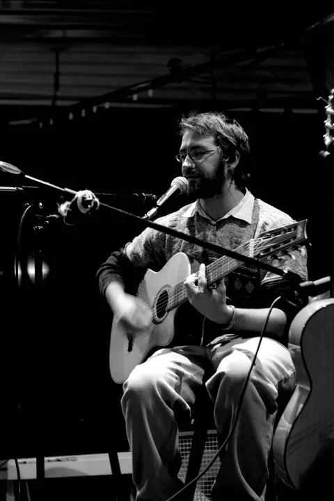 En noir et blanc, un des deux musiciens chante assis, guitare en main, devant un micro.