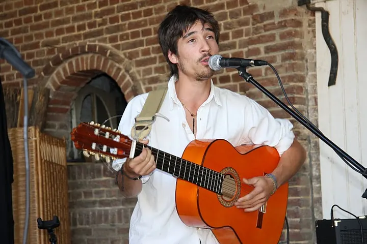 Devant un mur de briques, un musicien en chemise blanche chante avec une guitare orange.