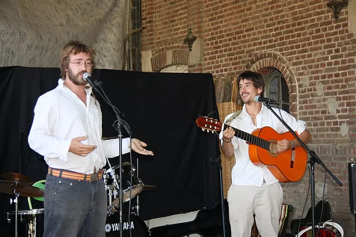 Le duo en concert dans une cour en briques : l'un chante les bras ouverts, l'autre joue de la guitare.